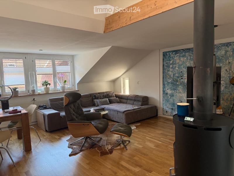 Modern living room with wooden floors, large windows, grey sectional sofa, leather armchair, small table, and a wood-burning stove.