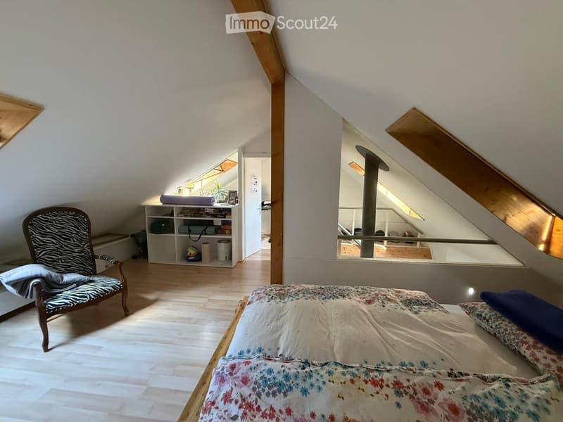 Cozy attic bedroom with wooden floors, slanted ceilings, a bed with floral bedding, a zebra-print armchair, a small shelf with books and decorative items, and a small table with a lamp.
