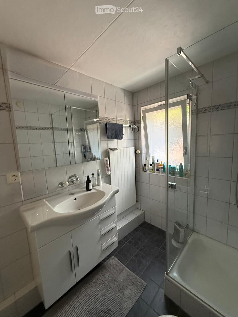 Modern bathroom with a white sink, mirror, shower, and various toiletries on the counter. The space features tiled walls and a dark tiled floor.