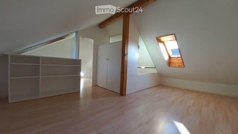 Bright attic room with wooden flooring, exposed wooden beams, built-in shelf unit, and a white wardrobe. A skylight window allows natural light to enter.