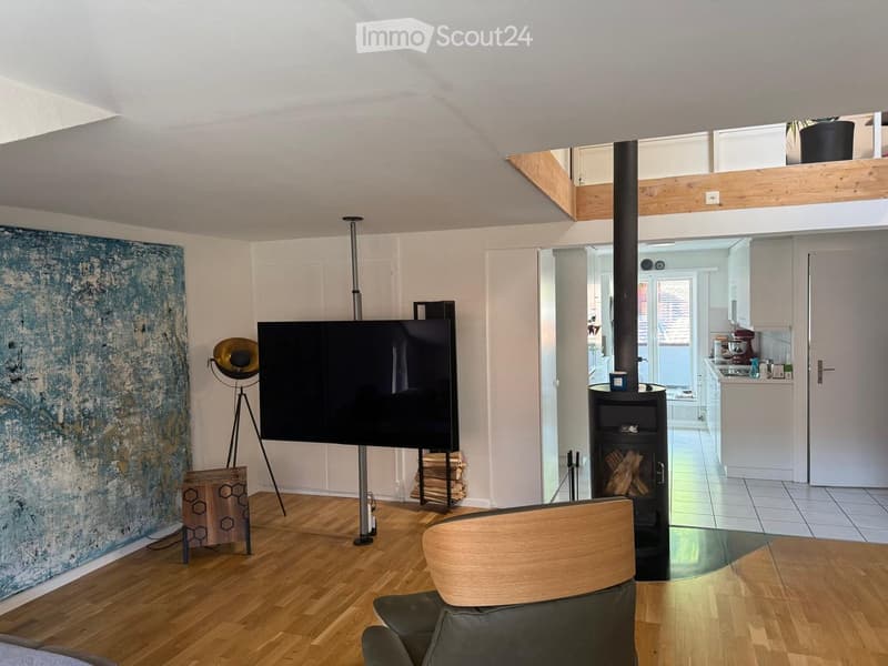 Modern living room with wooden flooring, a large television, a floor lamp, a wood stove, and an open kitchen area with white cabinets and appliances.