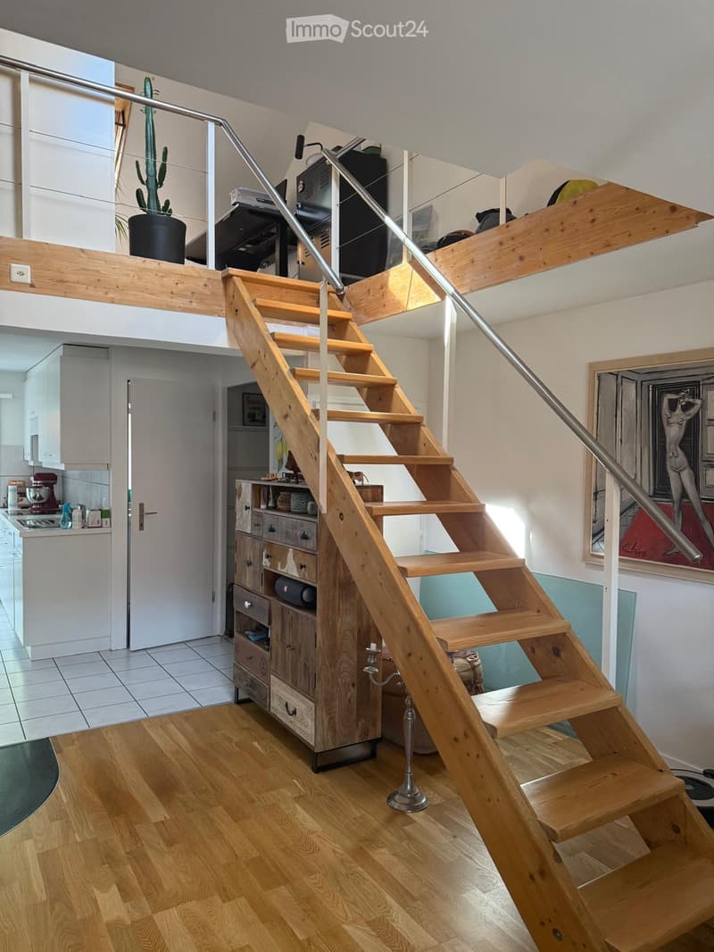 Modern hallway with wooden staircase, metal railing, and open loft area above. Features include a kitchen area with cabinets and appliances, a wooden cabinet with drawers, and a potted plant on the loft level.