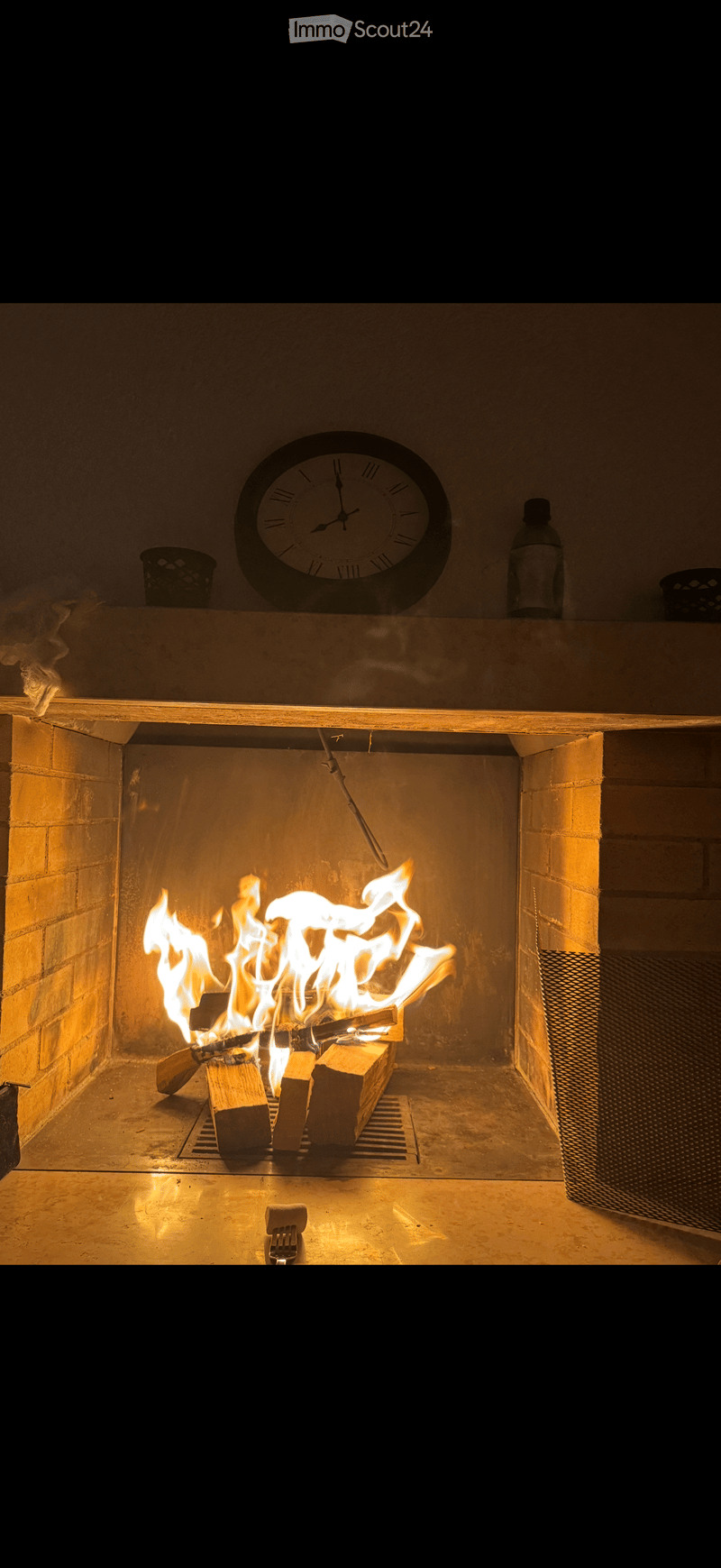 A lit fireplace with logs and flames, a clock above, and a bottle on the mantel.