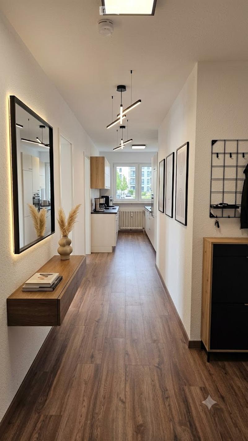Modern hallway with wooden flooring, recessed lighting, and a small console table with a vase and books. The hallway leads to a kitchen area with white cabinets and a window.