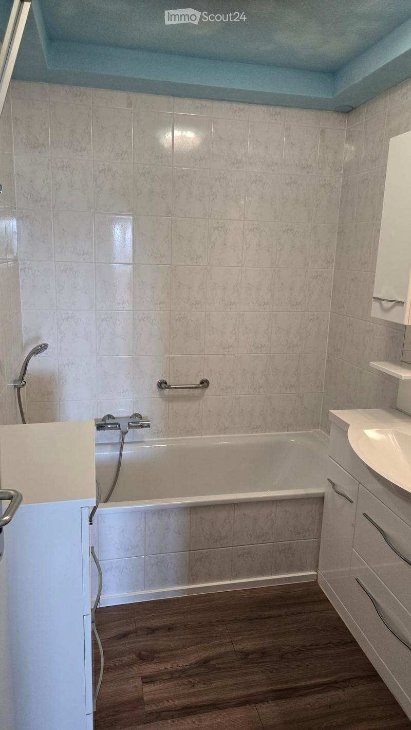 Modern bathroom with a bathtub, sink, and white cabinets. The walls are tiled with a marble-like pattern, and the floor is wooden.