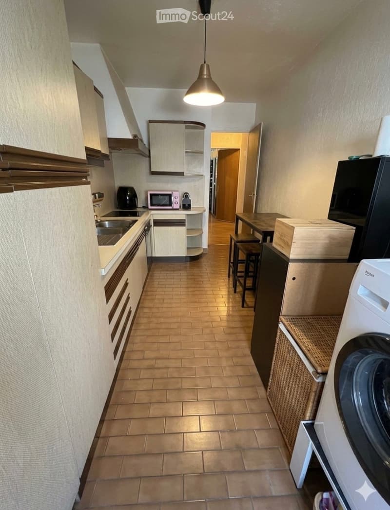 Modern kitchen with light wood cabinets, stainless steel sink, microwave, refrigerator, and washing machine. Includes a small dining area with a table and stools.