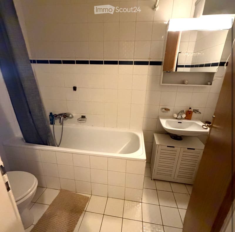 A small bathroom with white tiled walls and floor, featuring a bathtub, toilet, sink, mirror, and a small cabinet. A shower curtain is visible on the left side, and a bathroom rug is placed in front of the toilet.