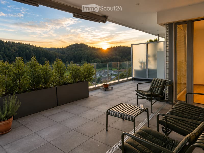 Balcony with tiled floor, glass railing, and scenic sunset view. Features lounge chairs, a small table, and potted plants.