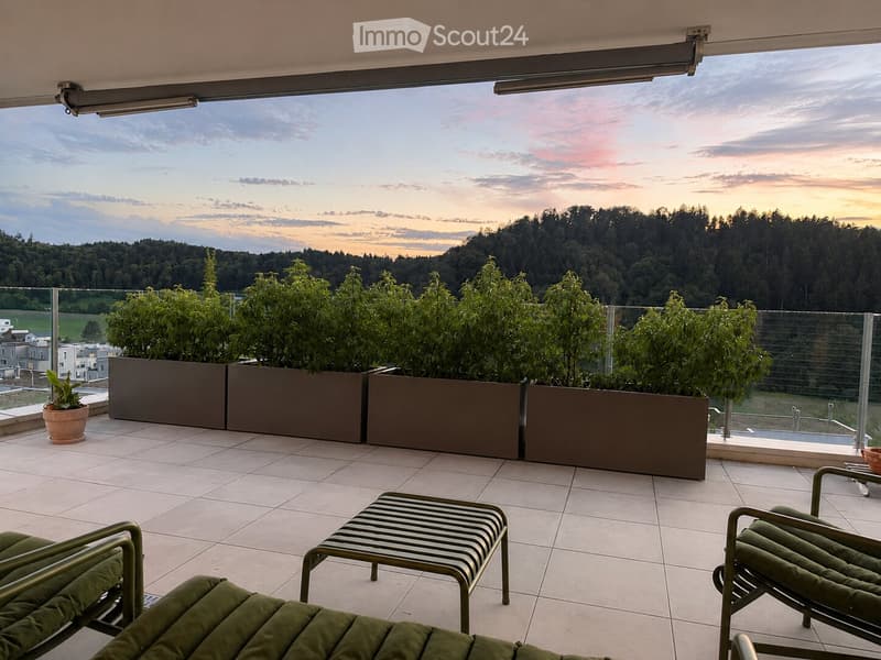 Balcony with tiled floor, green lounge chairs, small table, and potted plants, offering a scenic view of a forested area at sunset.