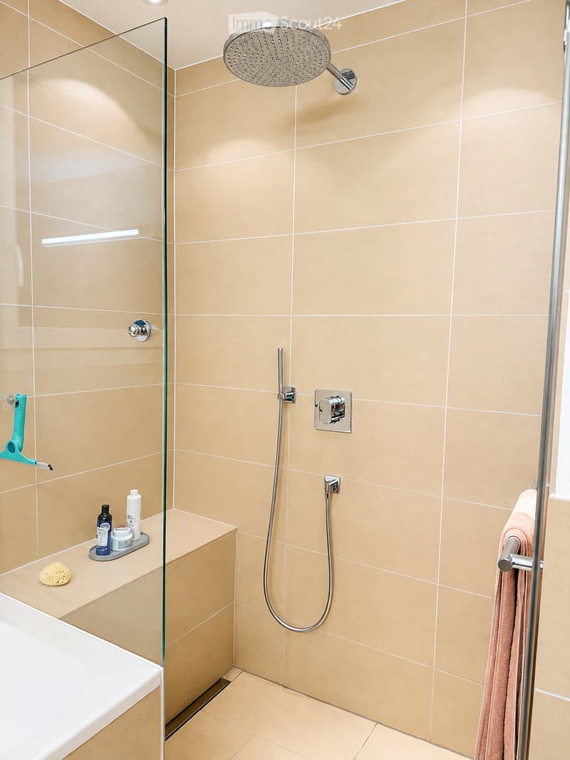 Modern bathroom with a glass-enclosed shower, a large showerhead, a towel hanging on a rack, a bath, and a shelf with various bathroom accessories.
