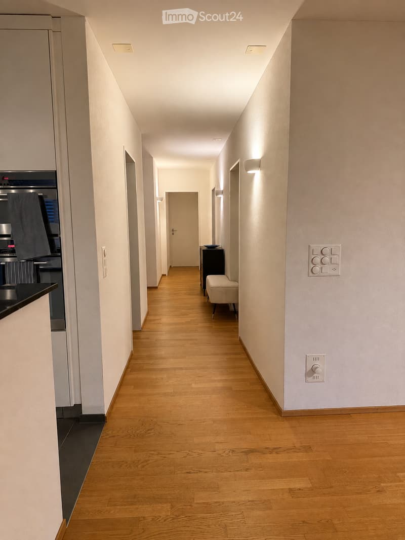 A long, narrow hallway with wooden flooring, white walls, and recessed lighting. Features include a small bench, a black cabinet, and a control panel with buttons and a knob on the wall.