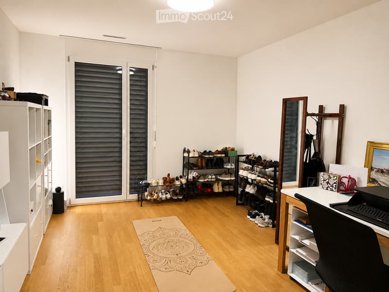 A well-lit room with wooden flooring, a large window with blinds, a desk with a chair, a shoe rack, a cabinet, and a mirror.