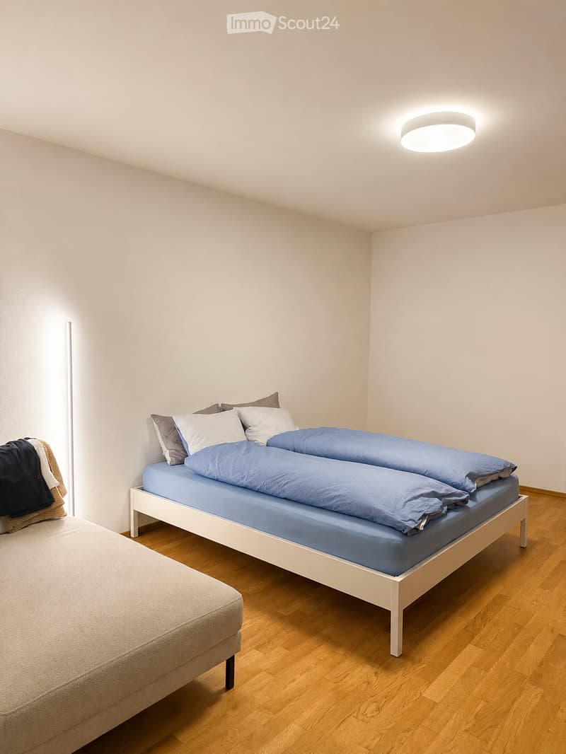A modern bedroom with a double bed, neatly made with blue bedding and pillows, a small sofa, and a wooden floor.