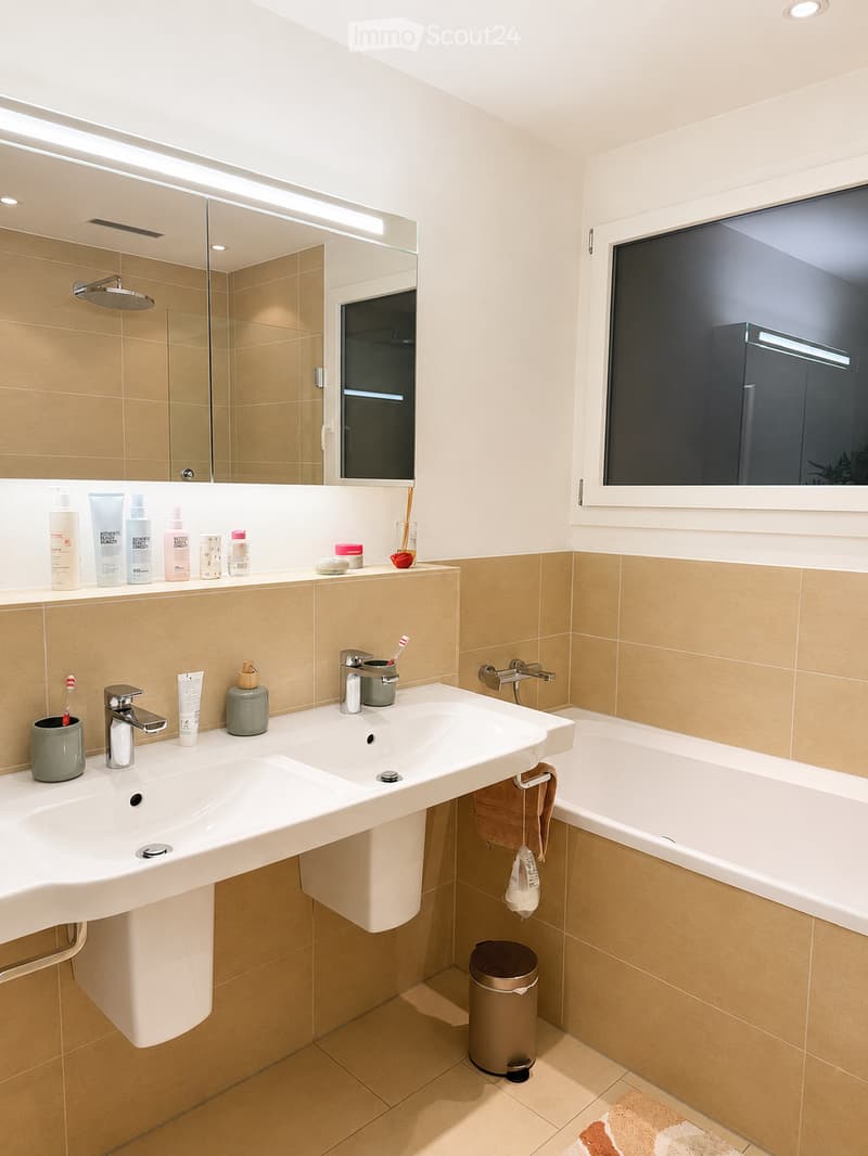 Modern bathroom with a double sink, bathtub, large mirror, and various toiletries neatly arranged on the countertop.