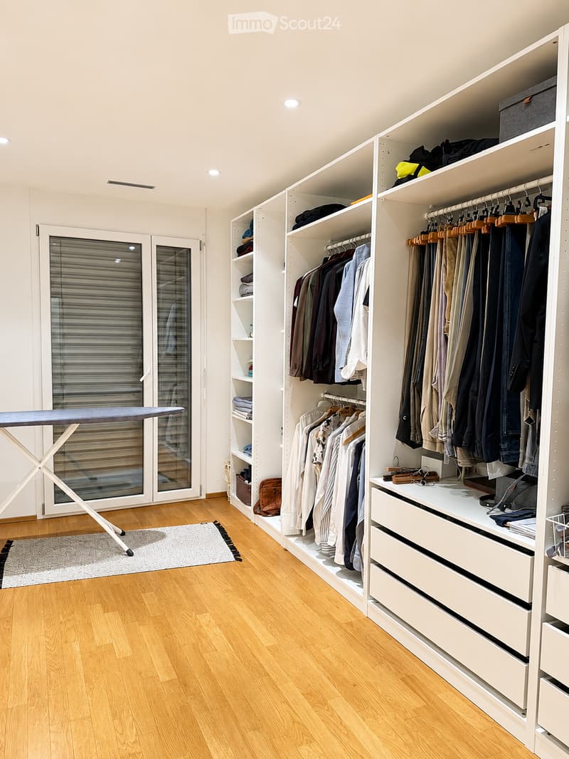 A well-organized walk-in closet with white built-in wardrobes, clothing racks, and storage drawers. An ironing board is positioned near the door, and a small rug lies on the wooden floor.