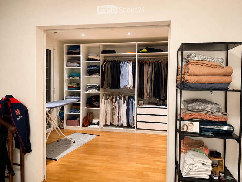 Well-organized walk-in closet with wooden flooring, built-in wardrobe with hanging space and shelves, and a separate shelving unit with folded blankets and pillows.