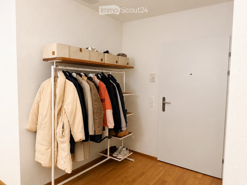 A modern, minimalist walk-in closet with a wooden floor, white walls, and a white wardrobe with hanging clothes and shelves holding boxes and folded garments.