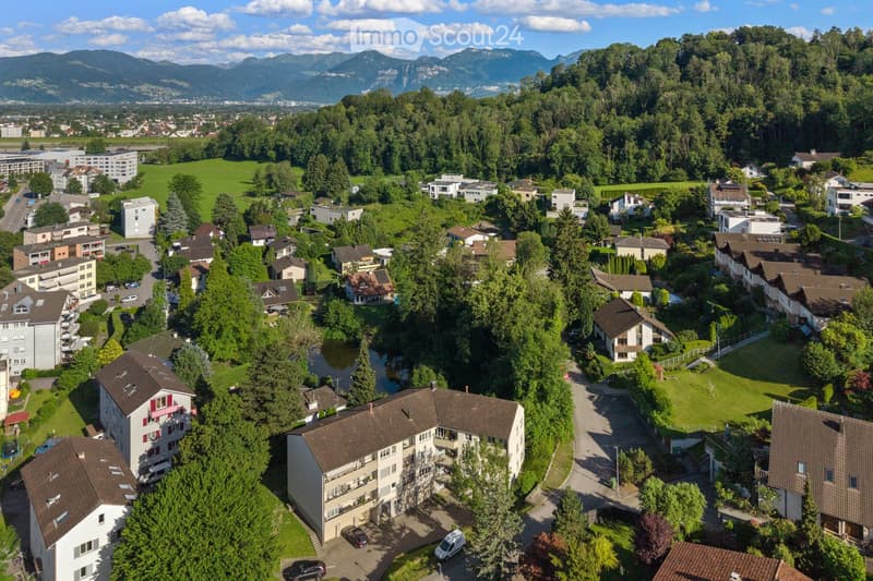 Aerial view of a suburban neighborhood with multiple residential buildings, surrounded by greenery and a river, with distant mountains in the background.