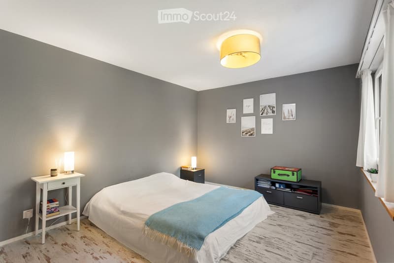 Modern bedroom with a double bed, nightstand, small table, and wall-mounted picture frames. The room features grey walls, wooden flooring, and a ceiling light fixture.