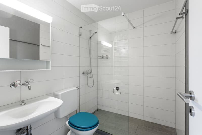 Modern bathroom with white tiles, a sink, toilet with a blue seat, a glass-enclosed shower, and a mirror with built-in storage.