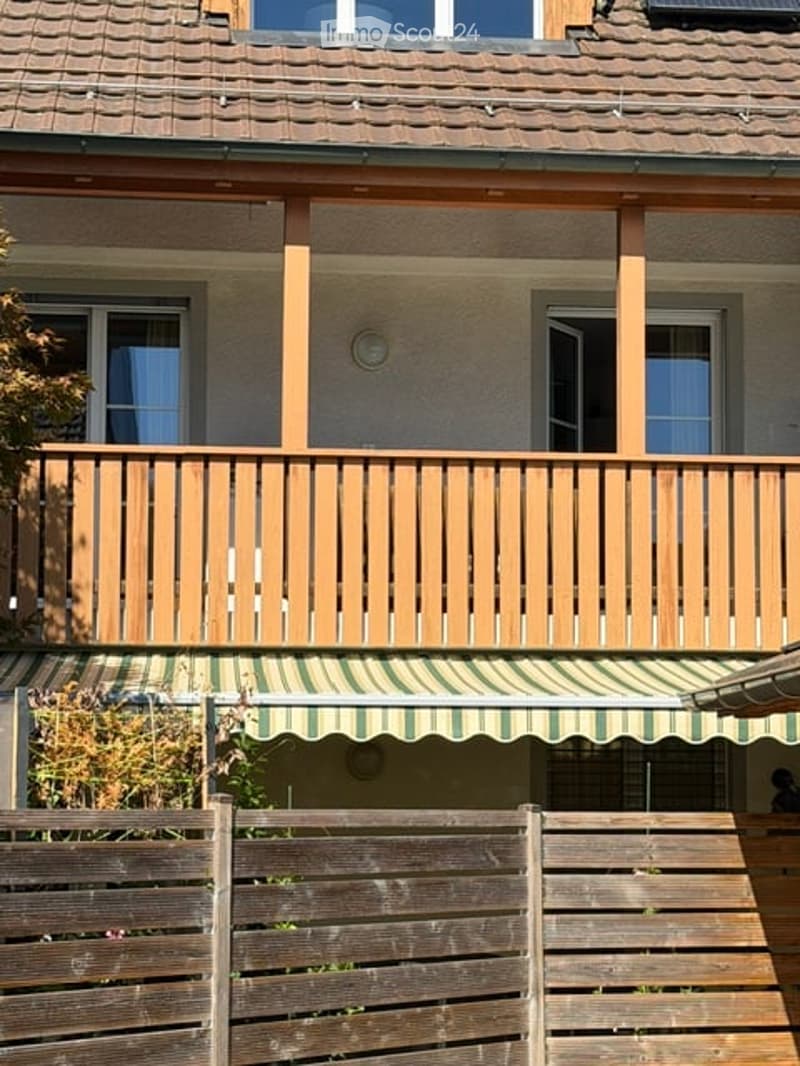 Two-story house with a wooden balcony and green striped awning. The balcony has wooden railings and is supported by wooden beams. The house has multiple windows and a wooden fence in the foreground.