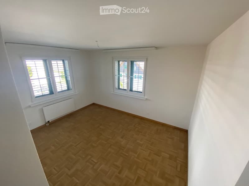 Empty room with wooden flooring, white walls, and two windows with blinds. Radiator visible under one window.