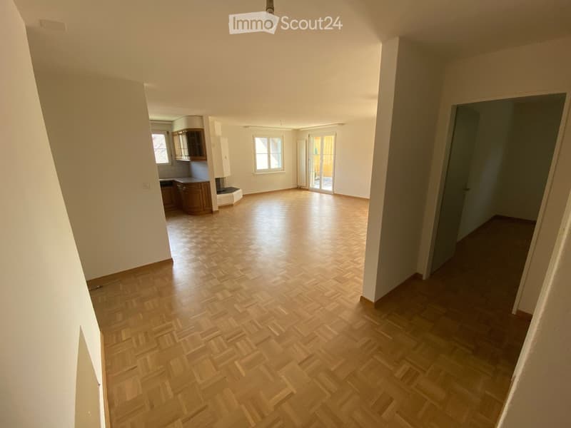Empty room with wooden flooring and white walls, featuring a doorway to another room and a small kitchen area visible in the background.