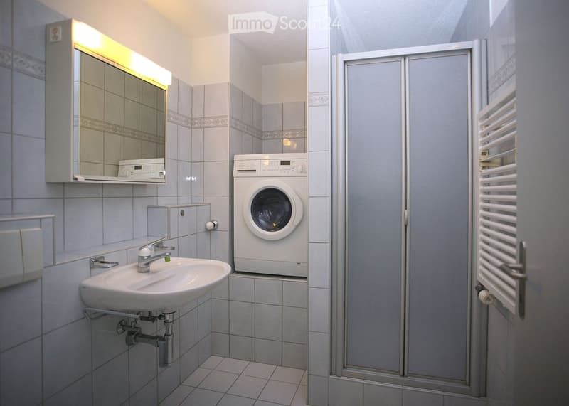 Modern bathroom with a sink, mirror, washing machine, and radiator. The space features tiled walls and a glass shower enclosure.