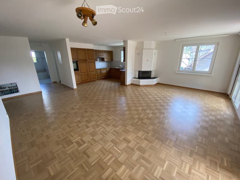 Empty room with wooden herringbone flooring, white walls, and a fireplace. The room has large windows and a visible kitchen area with wooden cabinets in the background.