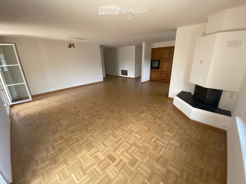 Empty room with wooden herringbone flooring, white walls, and a fireplace with a black insert. The room has large windows and visible ceiling lights.
