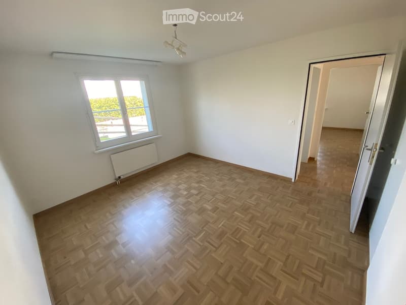 Empty room with wooden herringbone flooring, white walls, and a window with white frame. A radiator is visible below the window, and a ceiling light fixture is present.