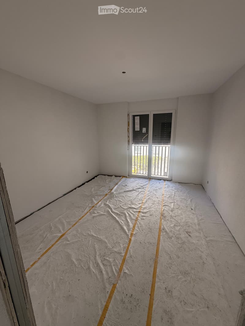 Empty room with bare walls and a protective plastic sheet covering the floor, featuring a sliding glass door leading to a balcony.