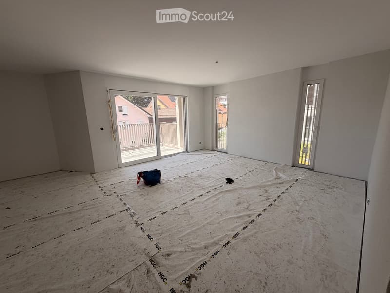 Empty room with bare walls and a protective floor covering, featuring large windows and a sliding door leading outside.