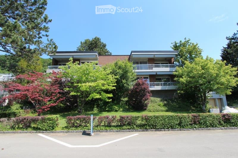 Two-story brick building with balconies, surrounded by lush greenery and trees.