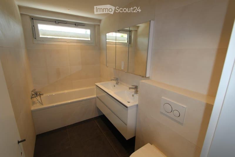 Modern bathroom with a bathtub, double sink, large mirror, and a window above the bathtub.