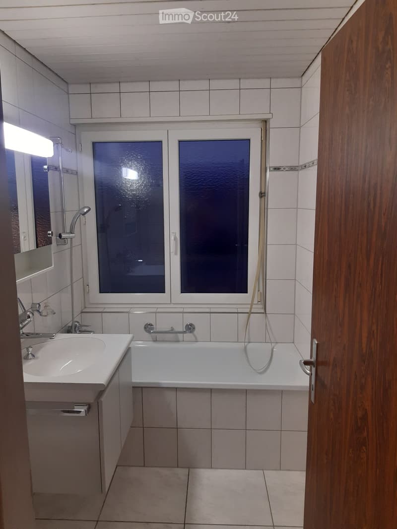 Bathroom with a white tiled wall, a bathtub, a sink, a showerhead, and a window with frosted glass panes.