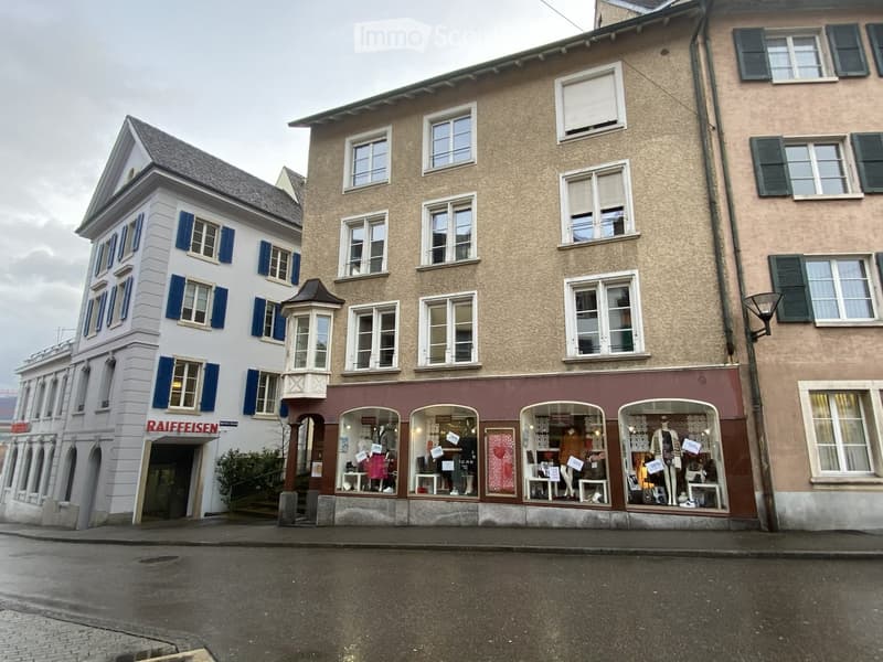 Three-story building with a storefront labeled "RAIFFEISEN" and display windows showing clothing and accessories. The building has a mix of white and beige facades with multiple windows and shutters.