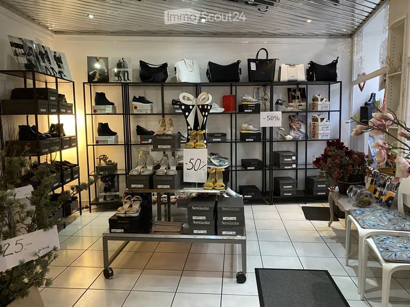 A well-lit retail space with shelves displaying shoes and handbags, a small table with boxes, and a decorative flower arrangement. The floor is tiled, and there are promotional signs visible.