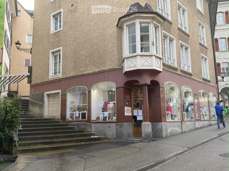 A multi-story building with a beige facade, white-framed windows, and a prominent bay window on the upper floor. The ground floor features a storefront with large glass windows displaying clothing and accessories, and a wooden double door entrance.