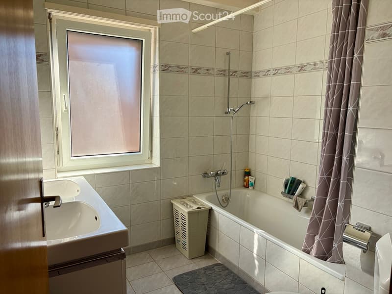 Bathroom with a bathtub, sink, showerhead, and shower curtain. Includes a toilet, toilet paper holder, and a small basket on the edge of the tub.