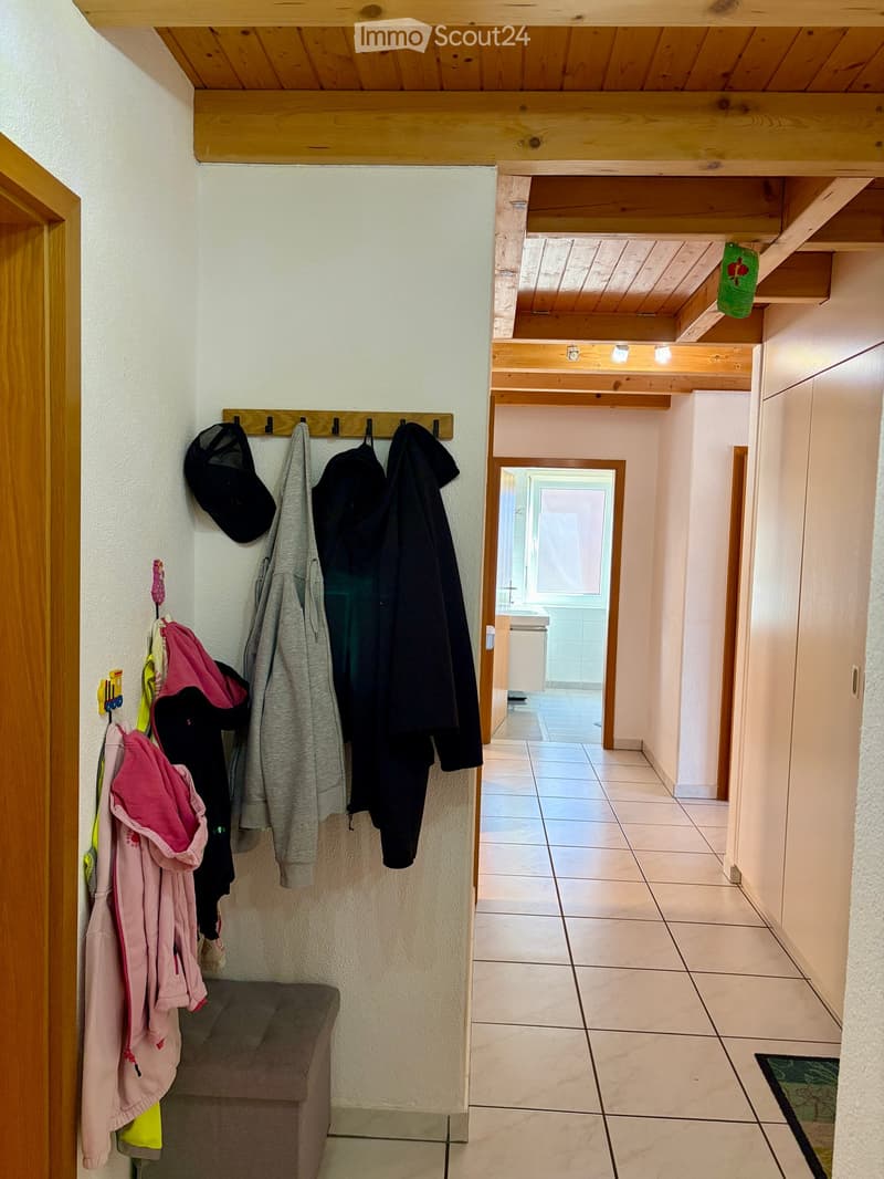 A hallway with wooden beams on the ceiling, tiled floor, and coats hanging on a rack on the left side. A small bench is visible near the rack, and a doorway leads to another room.