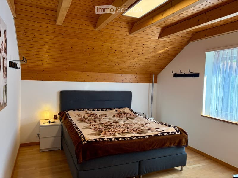 Cozy bedroom with wooden ceiling, slanted roof, large window with white curtains, and a neatly made bed with a patterned duvet and pillows.