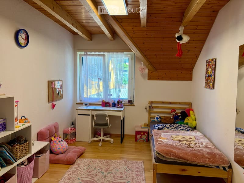 Cozy children's bedroom with wooden slanted ceiling, bed with pink bedding, desk with chair, shelf with toys, and a window with curtains.