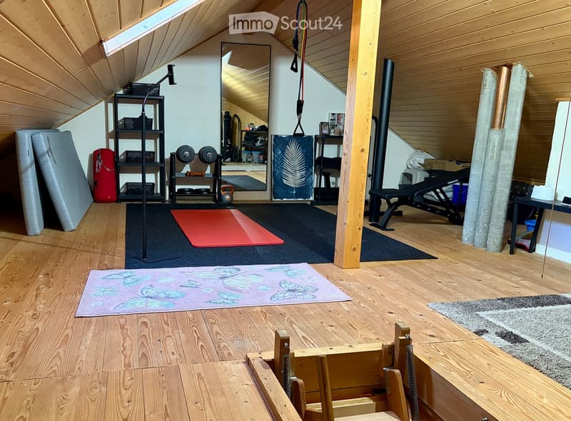 Well-lit attic space with wooden floors, exposed beams, and various fitness equipment including weights, a bench, exercise mats, and storage shelves.