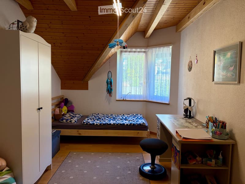 Cozy bedroom with wooden slanted ceiling, bed with blue and white checkered bedding, desk with chair, wardrobe, and dreamcatcher hanging on wall.