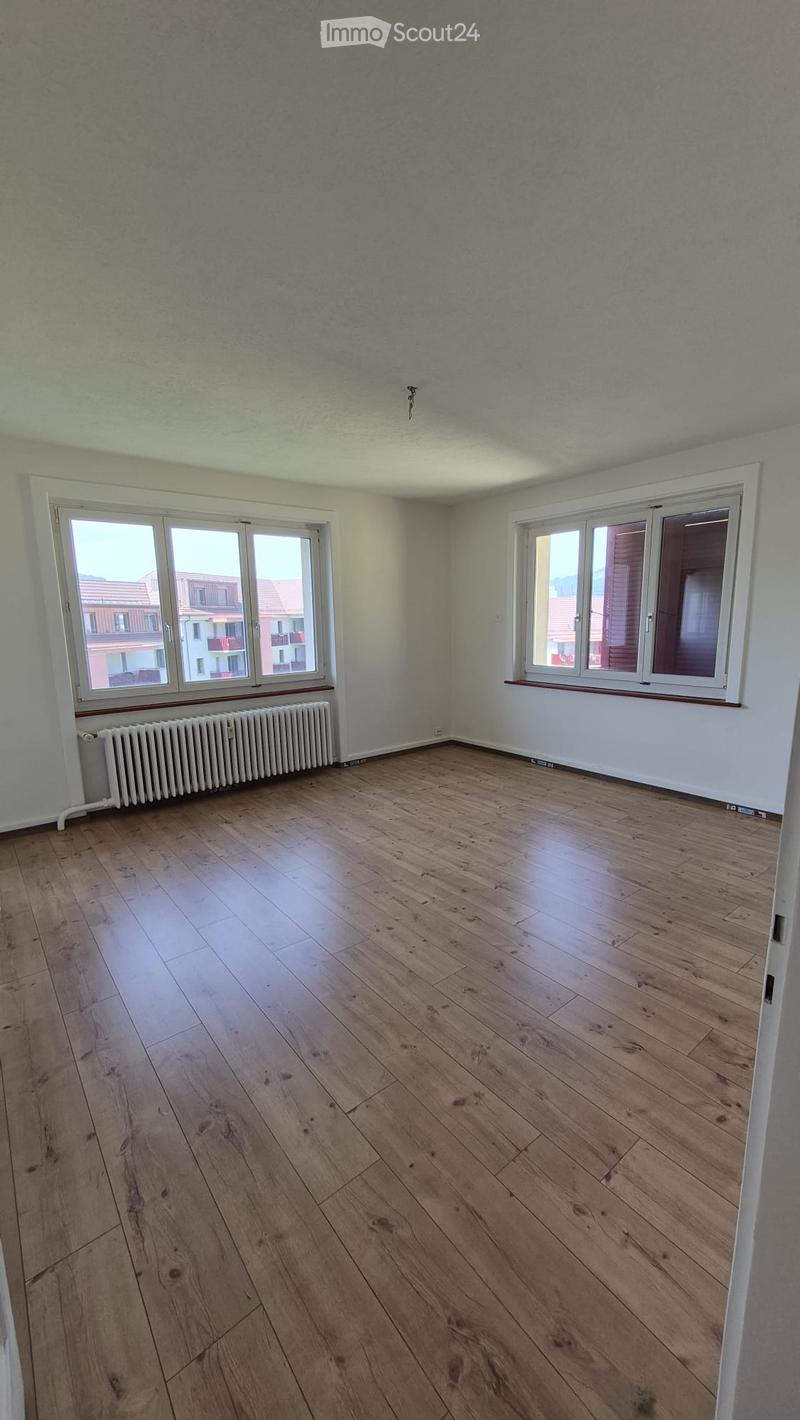 Empty room with wooden flooring, white walls, large windows, radiator, and visible electrical outlets.