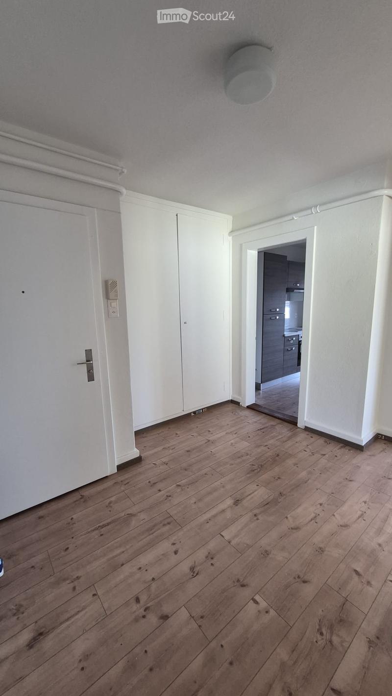 Empty hallway with wooden flooring, white walls, and a closed white door on the left. A doorway leads to another room with grey cabinets and a kitchen area visible in the background.