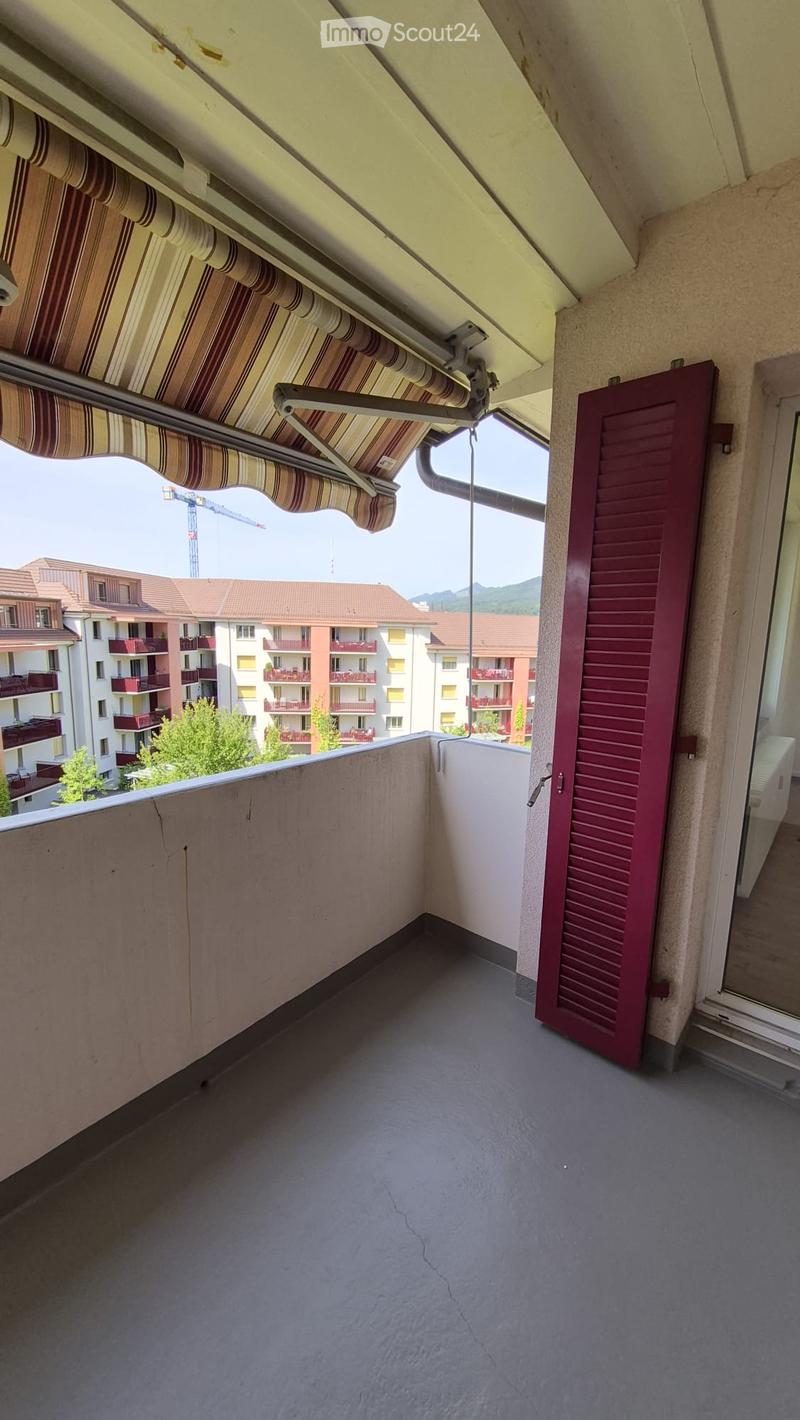 Balcony with grey concrete floor, red shutters, striped awning, and view of neighboring buildings and greenery.