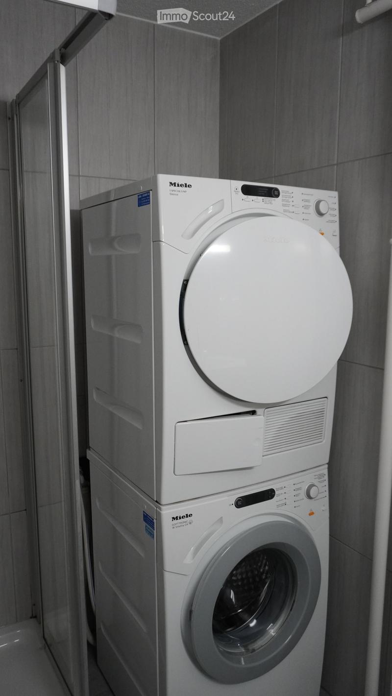 A modern bathroom with a stacked Miele washing machine and dryer unit, featuring sleek white finishes and a glass shower enclosure.