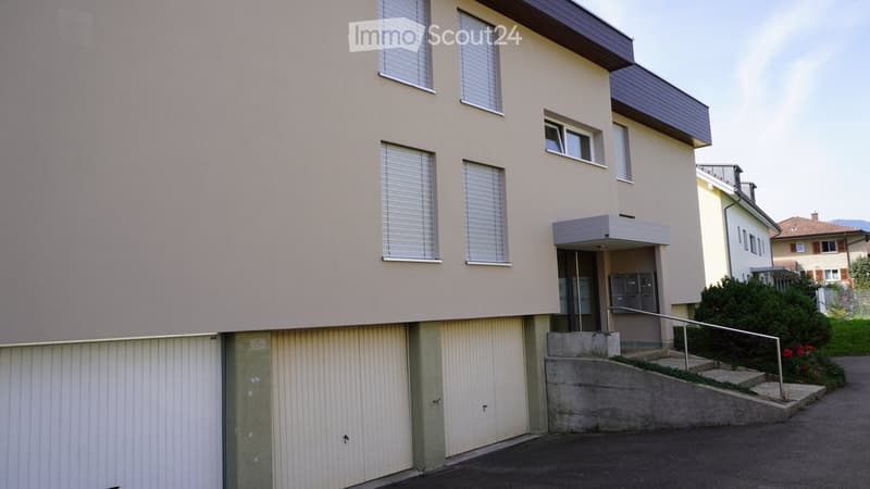 Two-story residential building with beige facade, multiple windows with blinds, a small entrance area with a staircase, and a garage on the left side.
